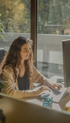Vrouw met krullend haar werkt aan een desktopcomputer bij grote ramen met herfstbomen buiten