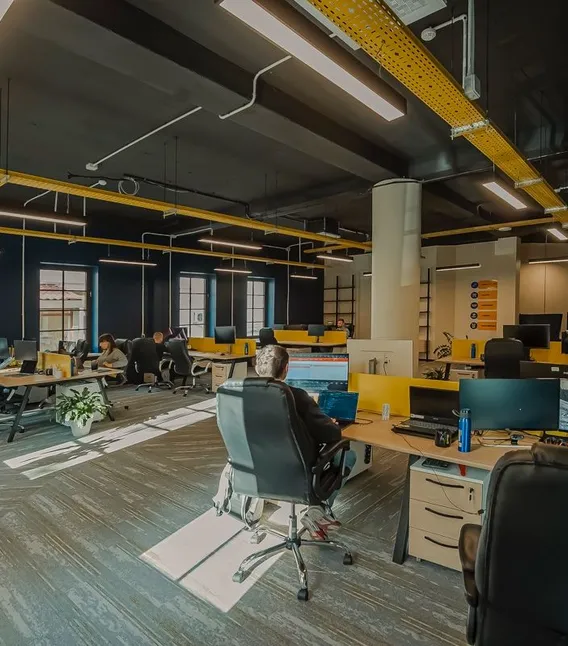 Een ruime open kantoorruimte met donkerblauwe muren en gele plafondaccenten, waar meerdere developers aan bureaus met monitoren werken.